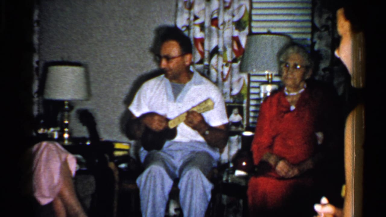 Man in a white shirt plays a small stringed instrument beside an elderly woman seated in a living room.