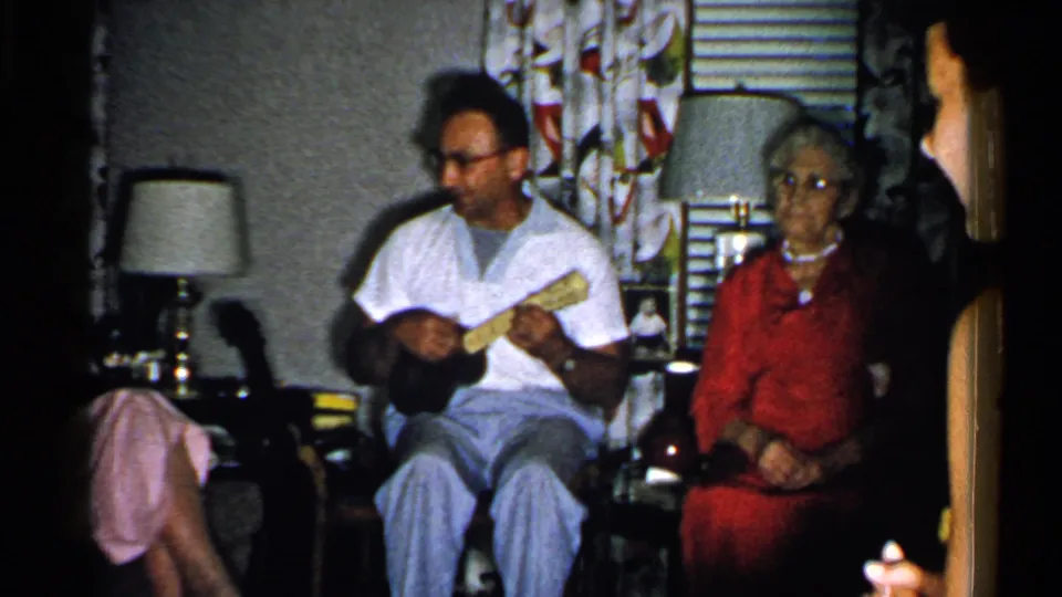 1952 New Hampshire home-movie frame of a man playing a small stringed instrument while an elderly woman in red sits nearby in a living room.