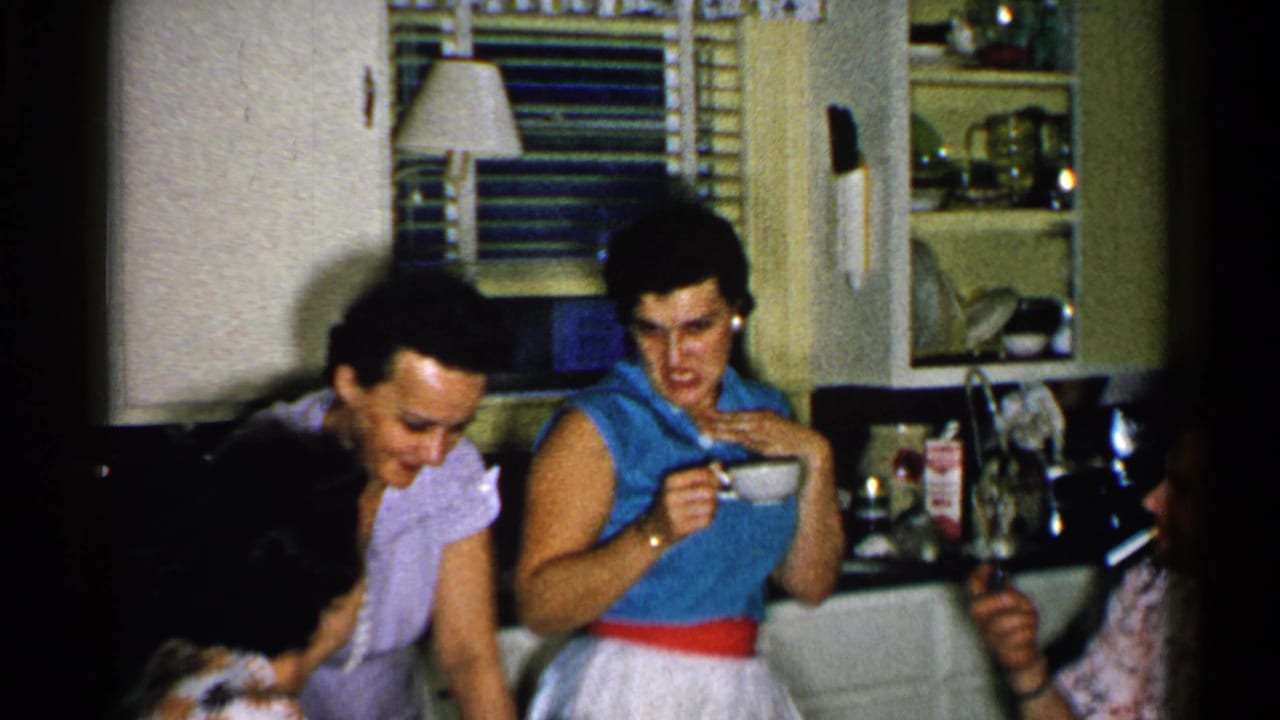 Women gather in a kitchen, with one woman sipping from a cup near shelves and window blinds.