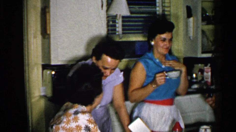 1952 New Hampshire home-movie frame of women in a compact kitchen, one holding a cup while others gather near the counter.