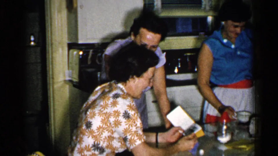 1952 New Hampshire domestic frame showing a woman holding an open album or booklet at a kitchen counter with other women nearby.