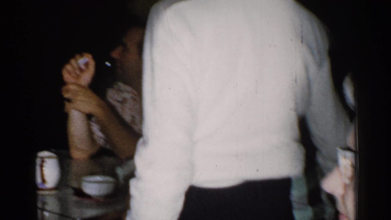 Adults seated at a table while a person in a white sweater stands in the foreground.
