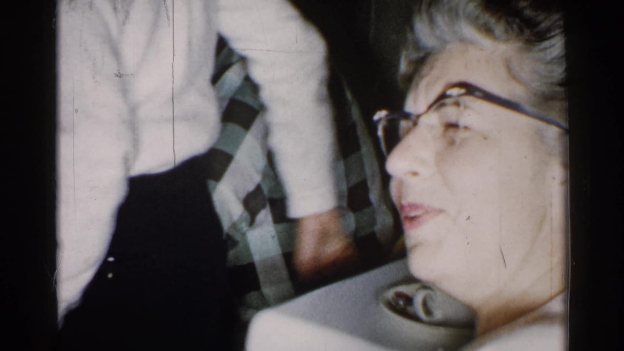 Older woman with glasses at right while a white-sweatered figure and patterned tie appear nearby.