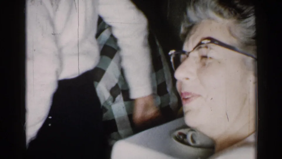 1952 New Hampshire interior frame with an older woman in glasses beside a standing figure in a white sweater and patterned tie.