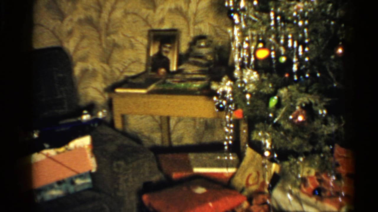 Living room corner with Christmas tree, wrapped gifts, couch, patterned wallpaper, and side table.