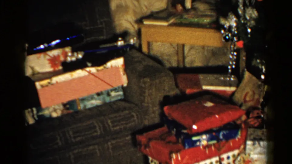 1952 New Hampshire home-movie frame showing a couch surrounded by wrapped Christmas presents, with a side table, wallpaper, and tree decorations nearby.