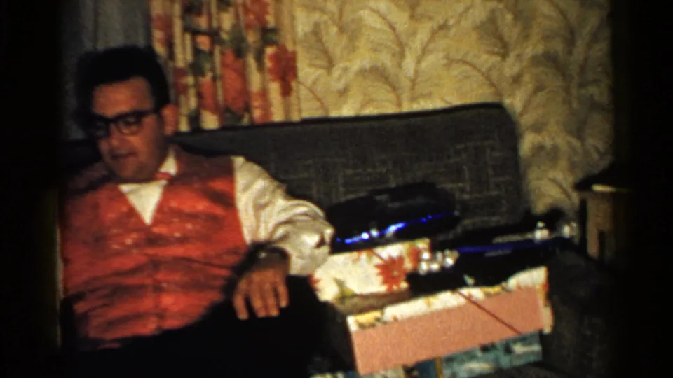 1952 New Hampshire home-movie frame showing a man in glasses and a festive red vest seated on a couch near wrapped Christmas presents.