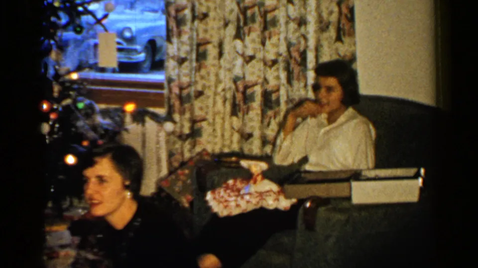 A 1952 New Hampshire holiday gathering frame with a seated woman, decorated tree, wrapped gifts, curtains, and a car visible through the window.