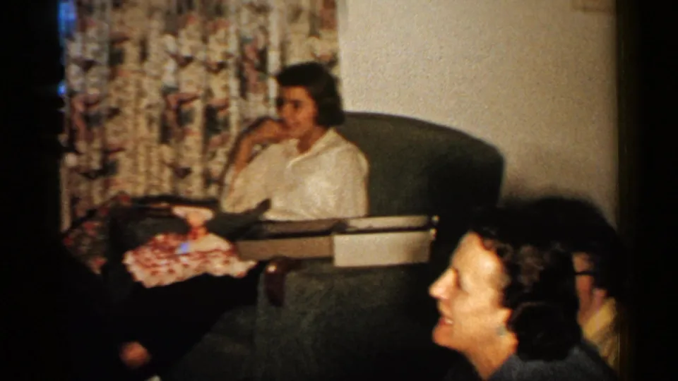1952 New Hampshire interior frame showing women in a living room with wrapped Christmas presents, patterned curtains, and upholstered seating.