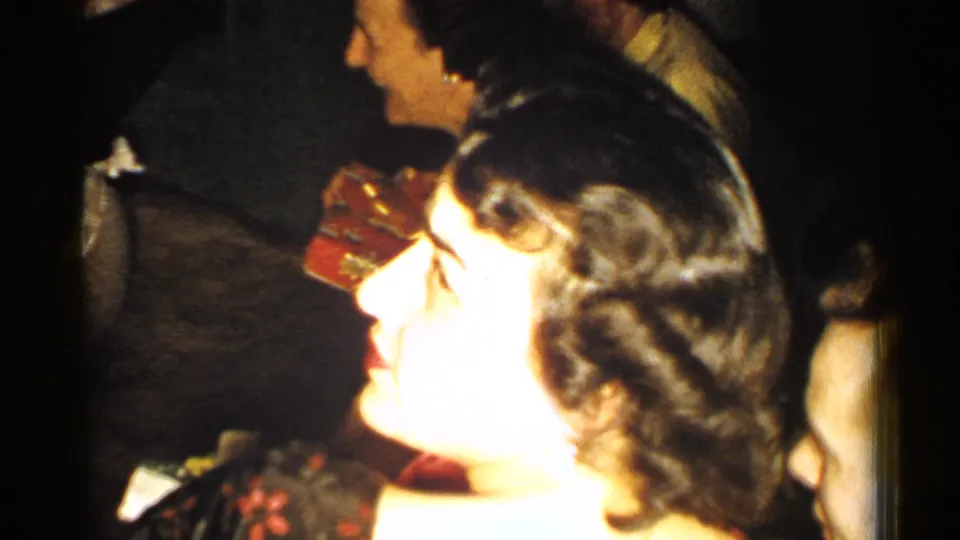1952 New Hampshire frame with a woman laughing in profile at an indoor family party, surrounded by other adults in soft 8mm footage.