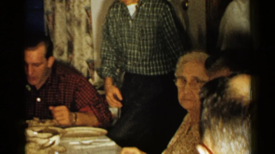 1952 New Hampshire home-movie frame showing adults around a dining table, with a plaid-shirted man standing and an older man seated nearby.