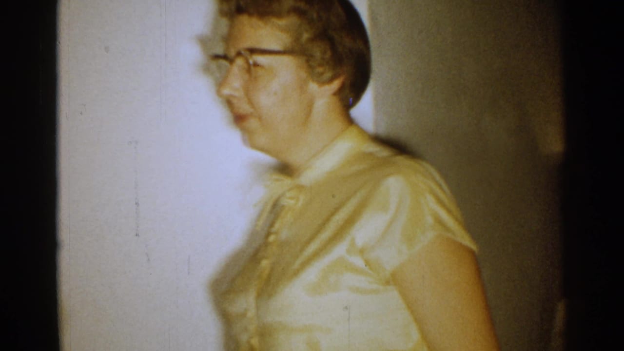 Woman in glasses and a yellow blouse walks past an interior wall and lamp.