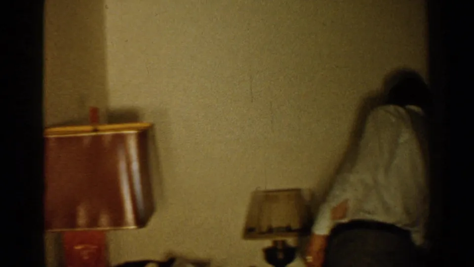 A man in a light shirt bends near a small table lamp in a private interior, with a larger brown-shaded lamp visible at left.