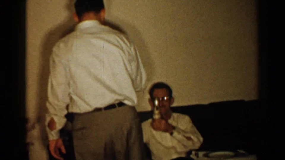 A standing man in a light shirt faces a seated man on a dark couch who holds a drinking glass in the 1952 New Hampshire interior scene.