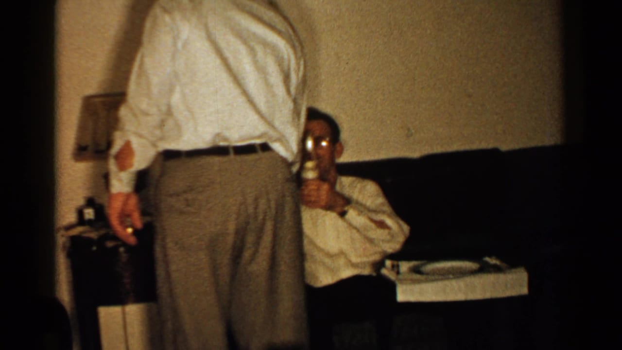Man standing with his back to the camera while another man sits on a couch holding a glass.
