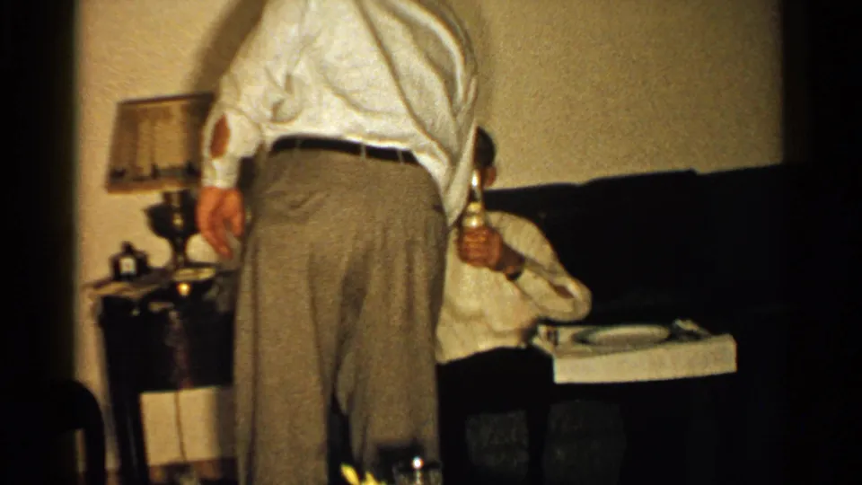 A 1952 New Hampshire interior frame shows two adult men near a couch, with a lamp and small table in view.