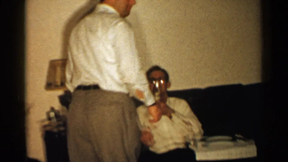 In a New Hampshire home-movie interior, a seated man smiles toward the camera while holding a glass near a couch.