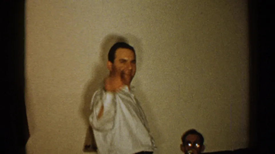 A smiling adult man in a white shirt turns near a plain wall in this 1952 New Hampshire home-movie frame.