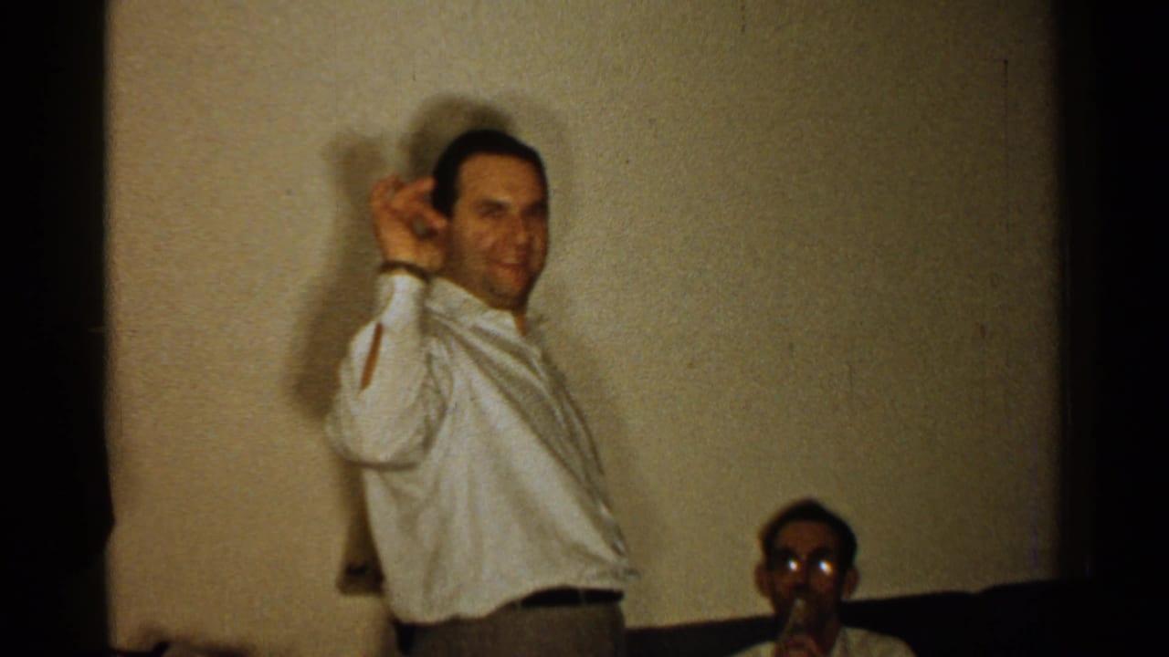 In 1952 New Hampshire home-movie footage, a man in a light shirt gestures beside a wall while another man sits partly visible below.