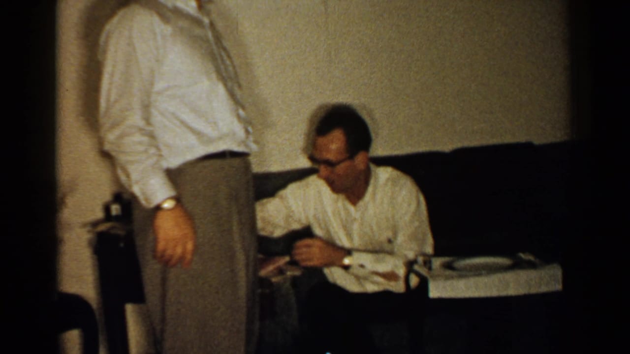 One man stands in front of a couch while another sits beside a small table in a living room.