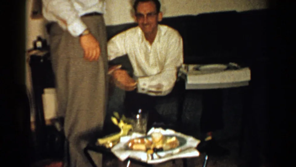 In a dim 1952 New Hampshire interior, a seated man faces the camera near snacks and a drink while another man stands partly in front.
