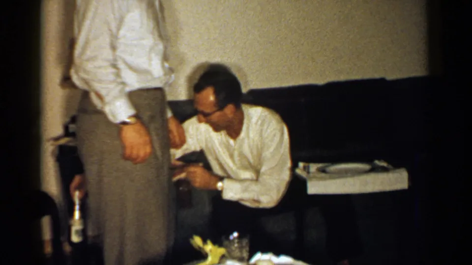 A 1952 New Hampshire home-movie frame shows a seated man handling a small object near a couch while another man stands in the foreground.