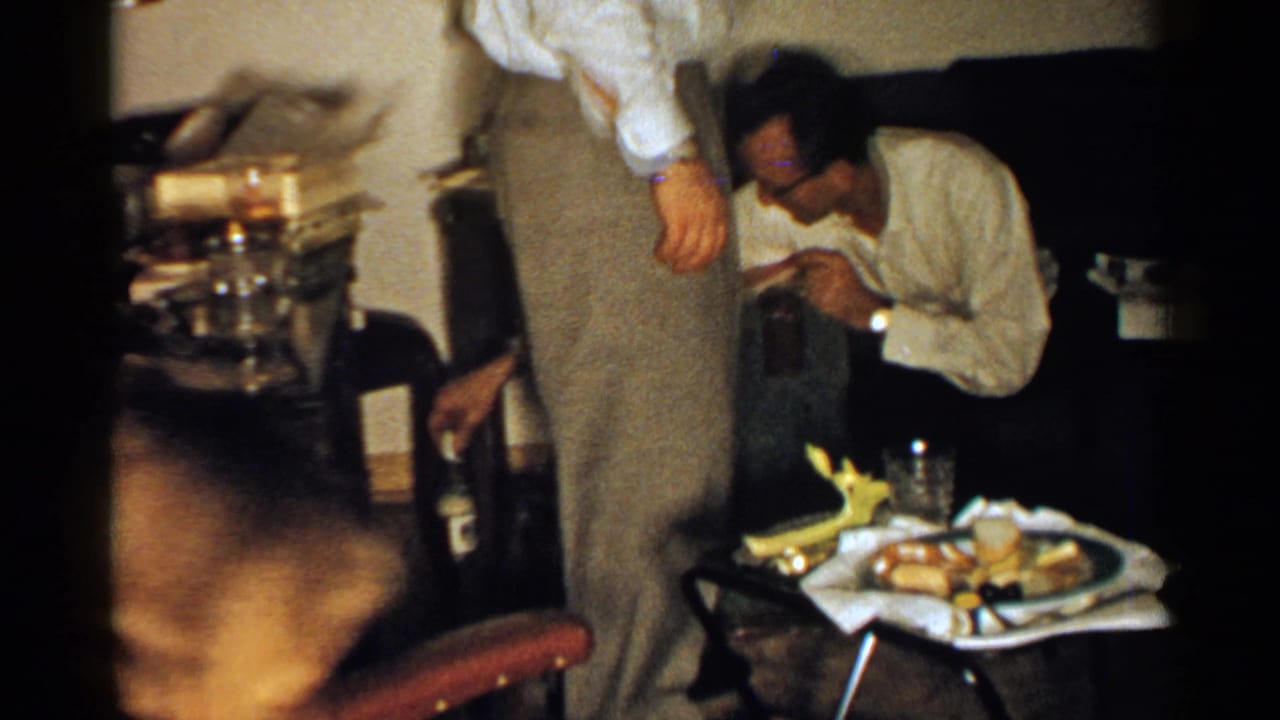 1952 New Hampshire 8mm frame showing adults in a dim home interior with food on a tray and a man seated near a dark couch.