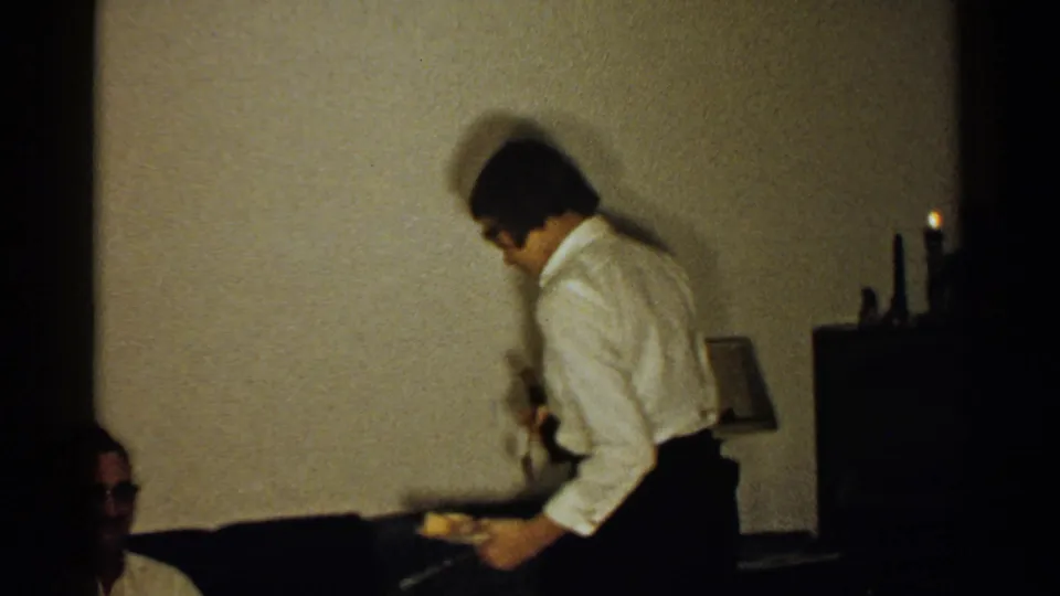 A woman in a white blouse and dark skirt carries a plate past a couch in a 1952 New Hampshire home interior.
