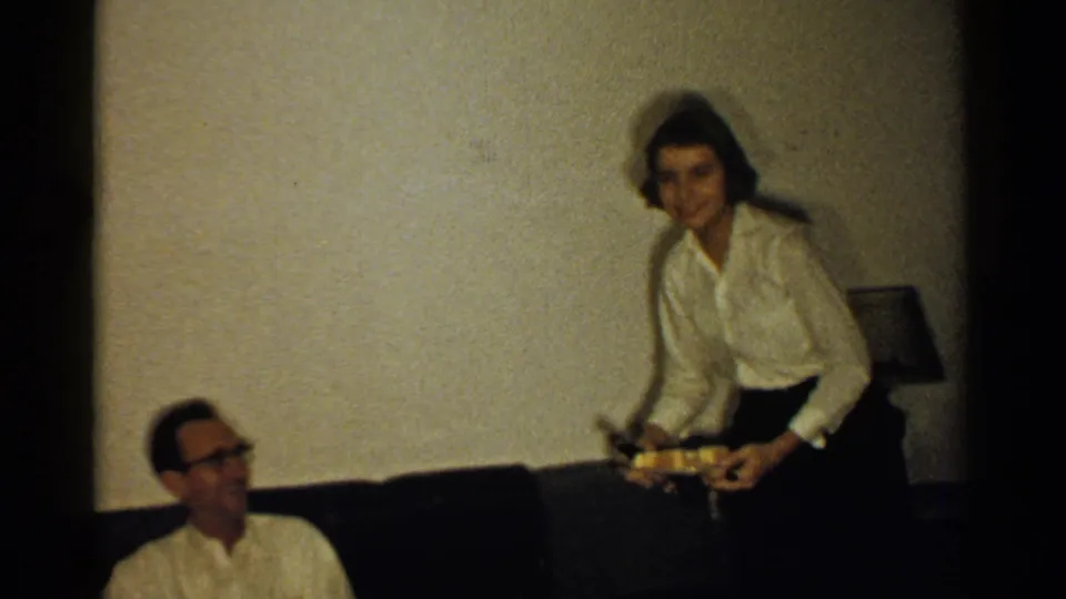 A woman in a white blouse holds a plate near a seated man in a dim 1952 New Hampshire living room.
