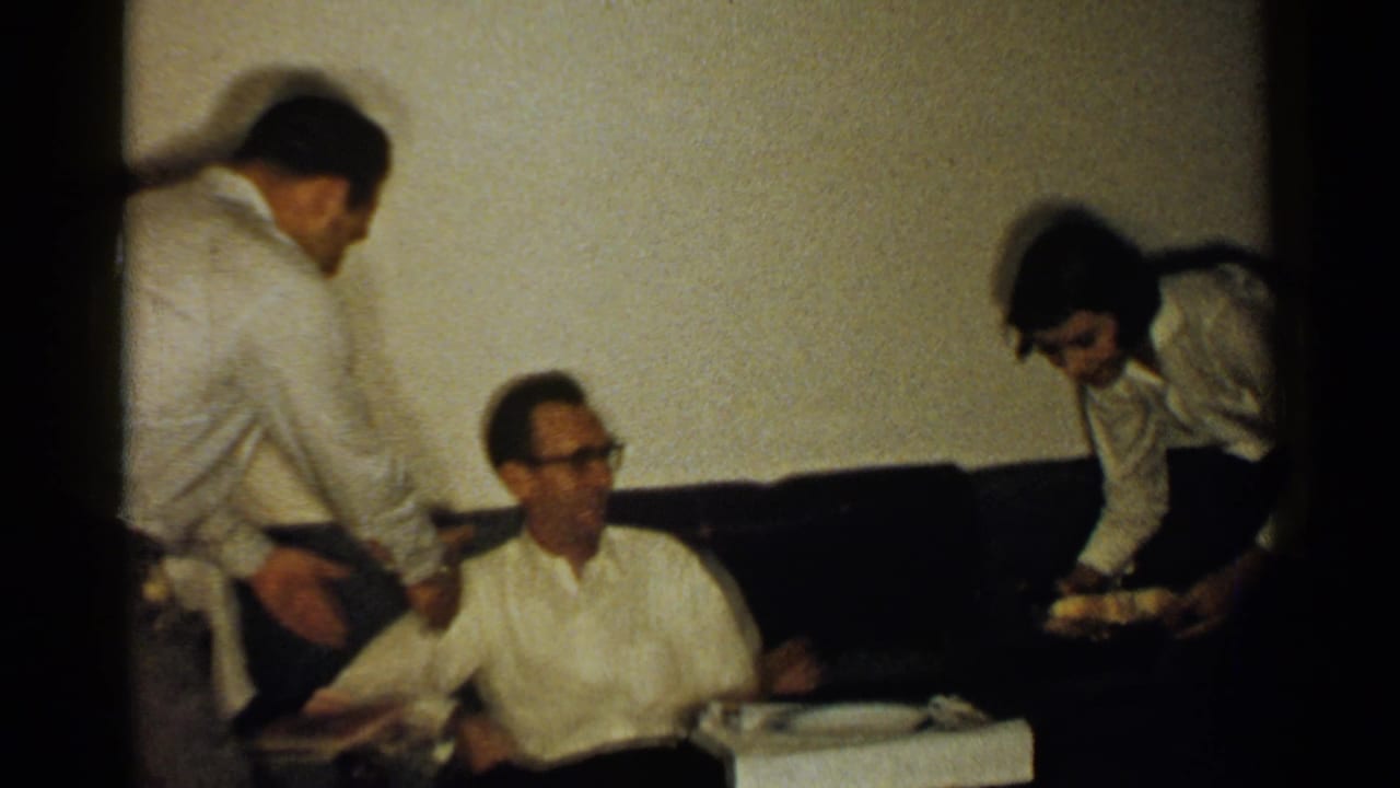 Three adults in a dim living room near a dark couch and low table.