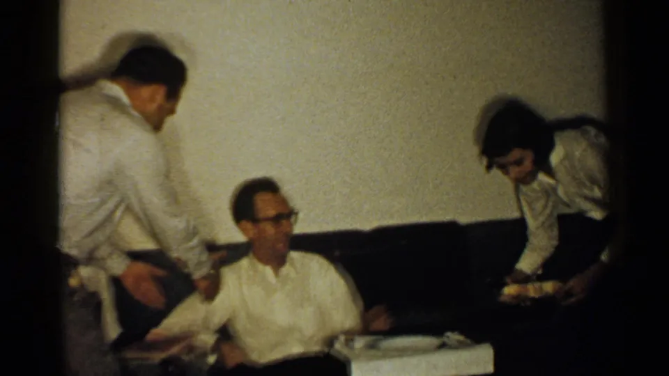 1952 New Hampshire home-movie frame showing three adults around a seated man on a dark couch in a dim living room.