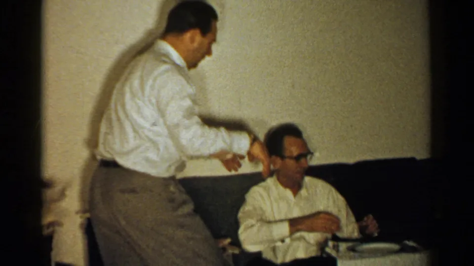 1952 New Hampshire living-room frame with a standing man leaning toward a seated man in glasses on a dark couch.