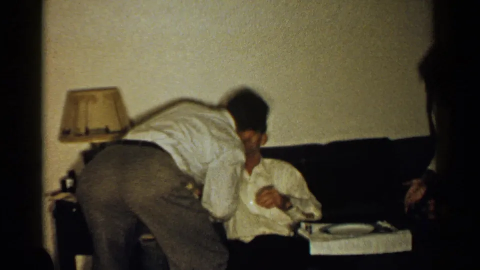 1952 New Hampshire home-movie frame showing a standing man bending in to kiss a seated man in a living room.