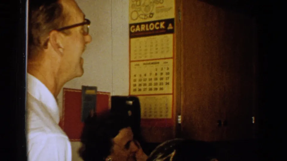 1952 New Hampshire home-movie frame showing an older man indoors near a Garlock calendar, with faces partly visible at the lower edge.