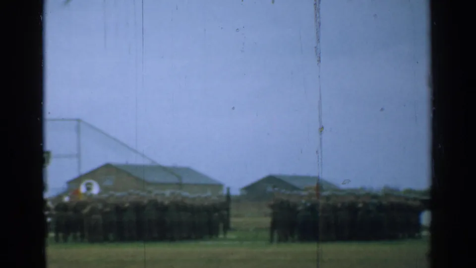 1952 New Hampshire 8mm frame showing distant soldiers in formation on a field, with low base buildings and window-like dark edges.