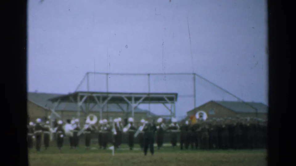 1952 New Hampshire home-movie frame showing a military band on a grassy field, soldiers in formation, base buildings, and a raised platform structure.
