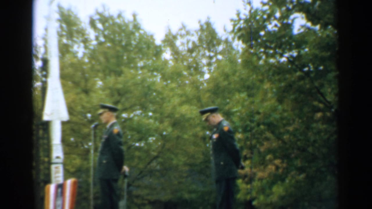 Two officers stand near microphones beside a white rocket model in a wooded outdoor setting.