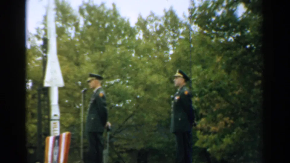 1952 New Hampshire 8mm still showing one officer at a microphone and another standing at attention near a white rocket model.