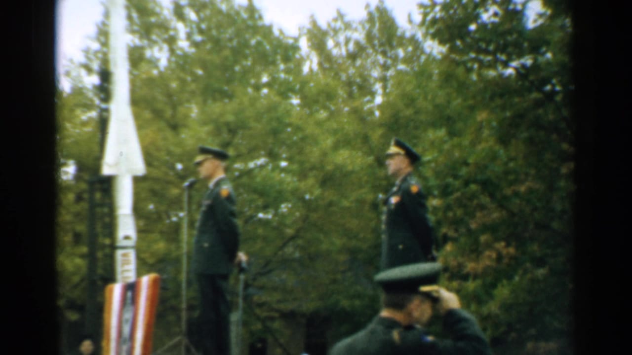 Uniformed soldiers stand outdoors near a white rocket-shaped monument and trees.