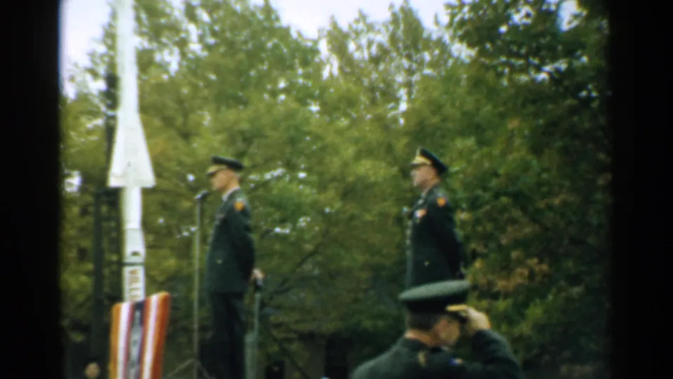 1952 New Hampshire home-movie frame showing uniformed soldiers at an outdoor ceremony beside a white rocket-shaped monument.