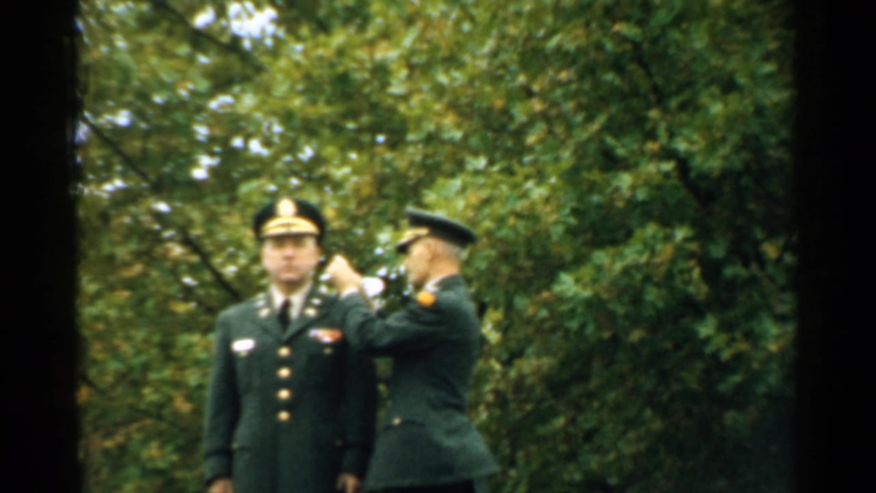 An officer stands at attention while another soldier reaches toward his shoulder or chest.