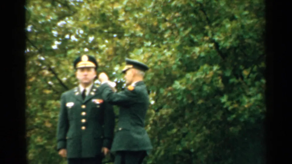 Mid-century New Hampshire home-movie frame showing an officer receiving a visible ceremonial adjustment or decoration outdoors.