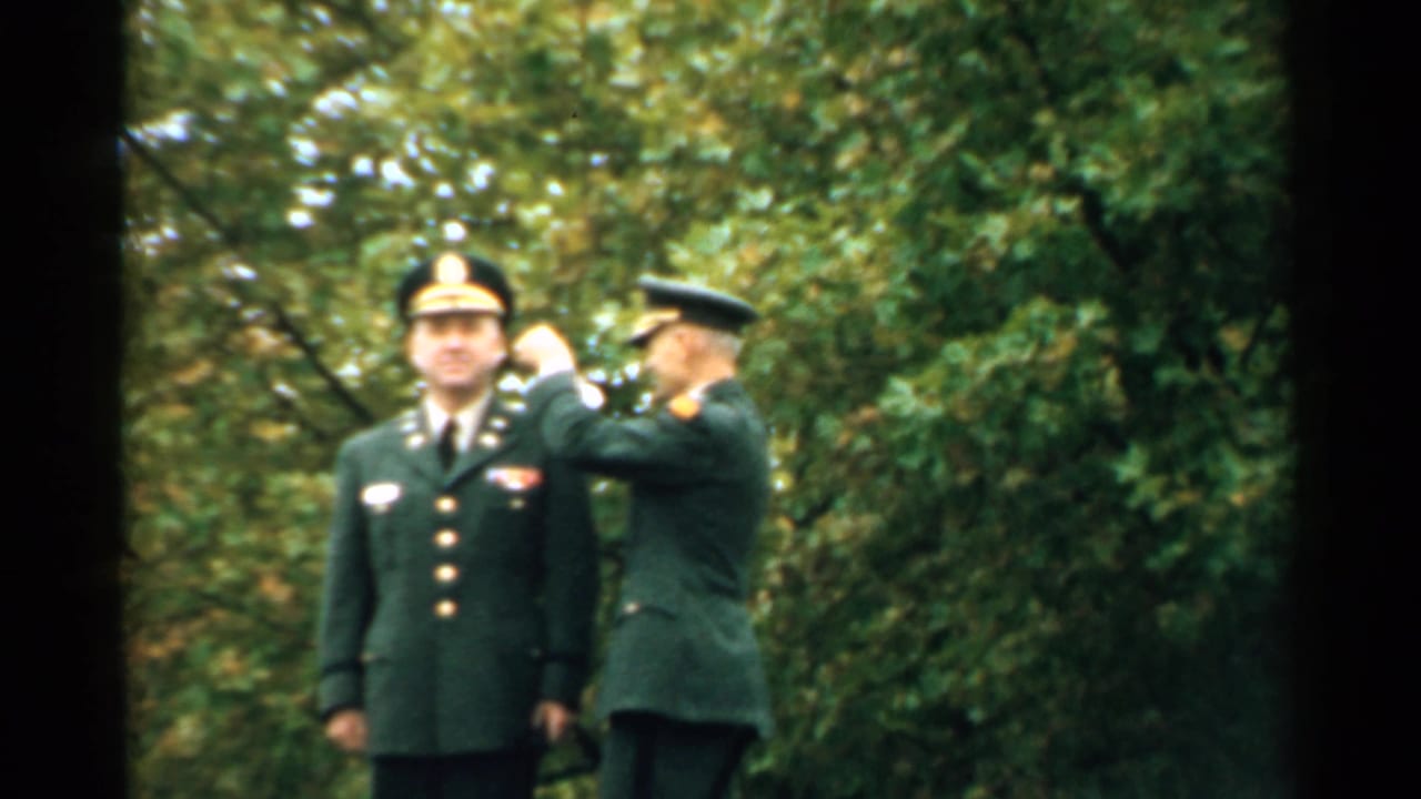 Two uniformed men stand outdoors as one pins an item on the other's lapel.