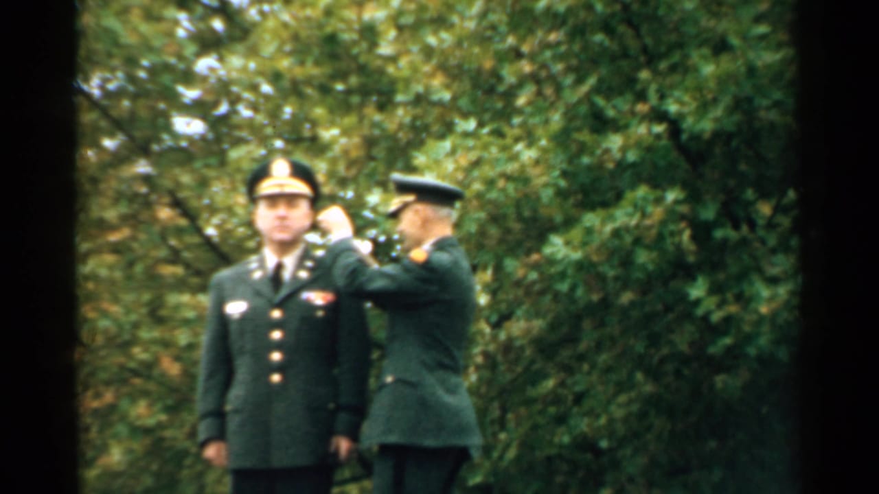 Uniformed officer reaches toward another man's lapel in a wooded outdoor setting.