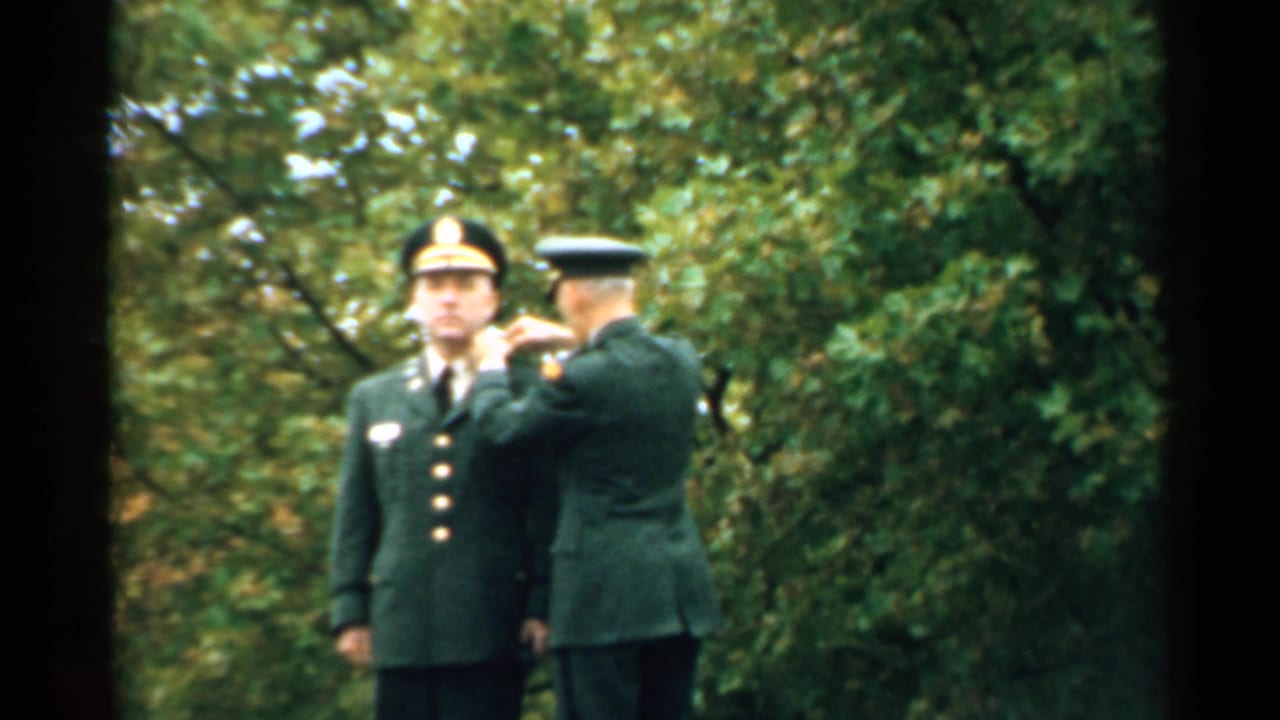 Officer pins a medal on a uniformed soldier standing at attention outdoors.