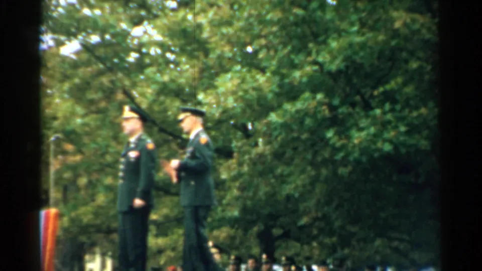 A certificate folder appears between two uniformed men during the 1952 outdoor military recognition scene.