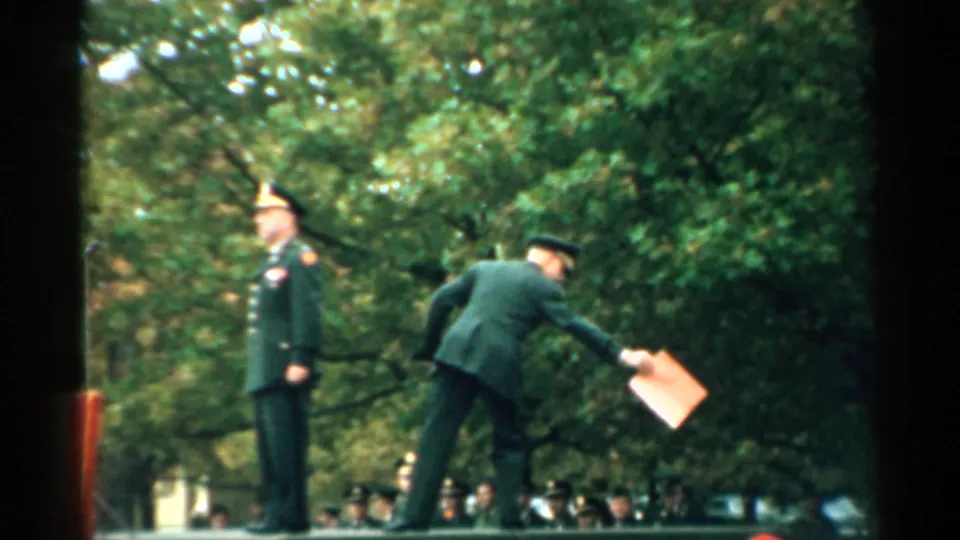1952 New Hampshire 8mm frame showing uniformed men at an outdoor military ceremony, with one officer standing and another passing a folder.