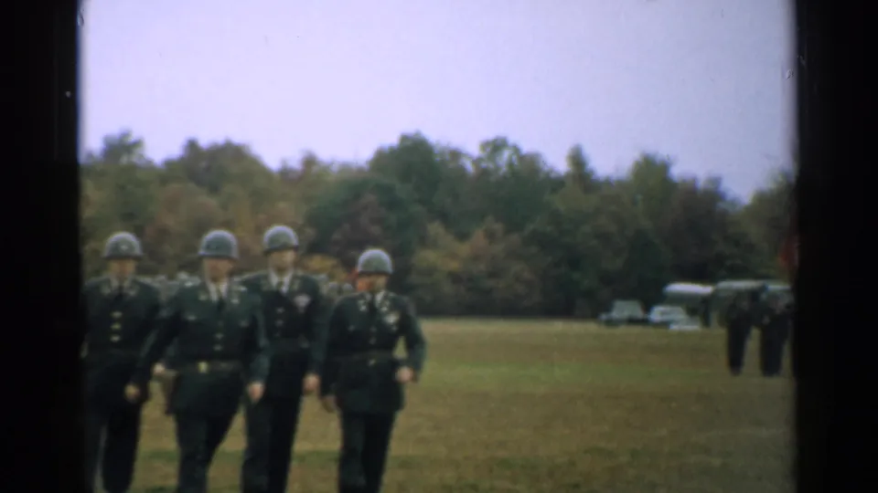 A 1952 New Hampshire home-movie frame shows soldiers in green uniforms and helmets marching across a grassy field.