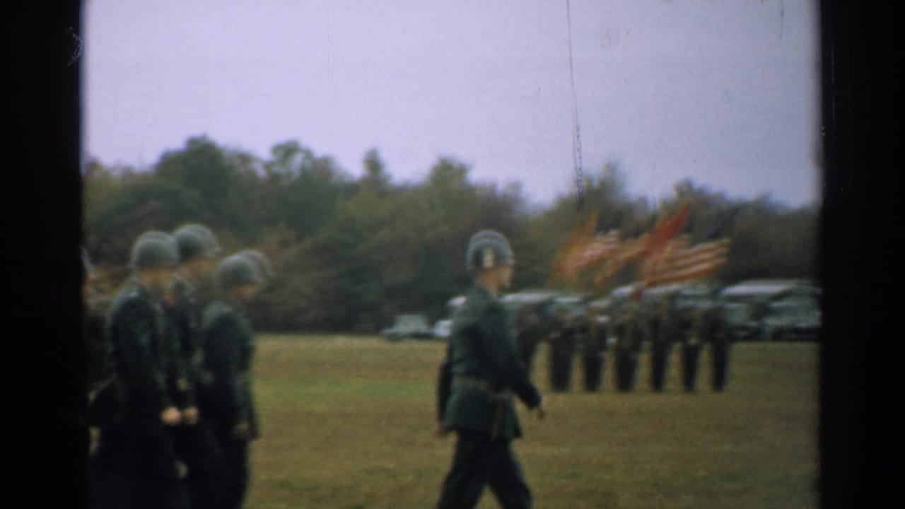 Soldiers in helmets march past a troop formation and American flags on a grassy field.