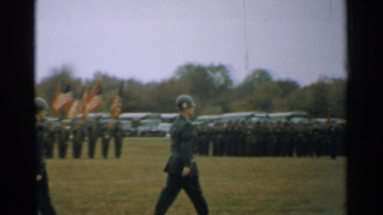 An officer walks across a field with soldiers, flags, and parked cars in the background.